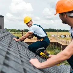 Deux couvreurs travaillent sur un toit en bardeaux gris, portant des casques de sécurité.